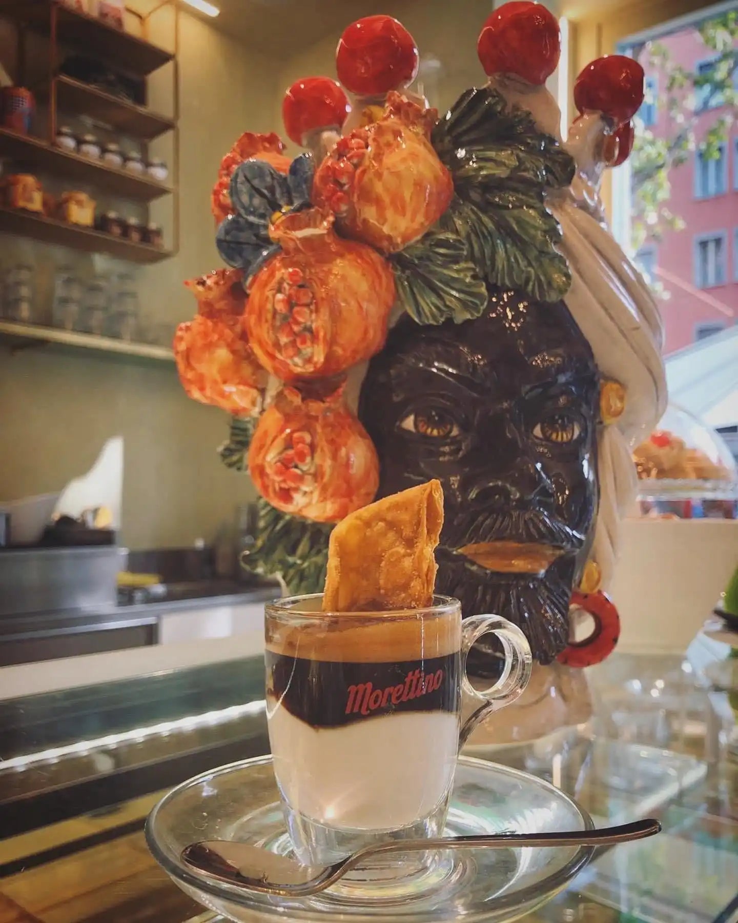 Vaso ornamentale in ceramica a forma di testa con rose arancioni e bacche rosse disposte sopra, esposto accanto a una bevanda di caffè stratificata.