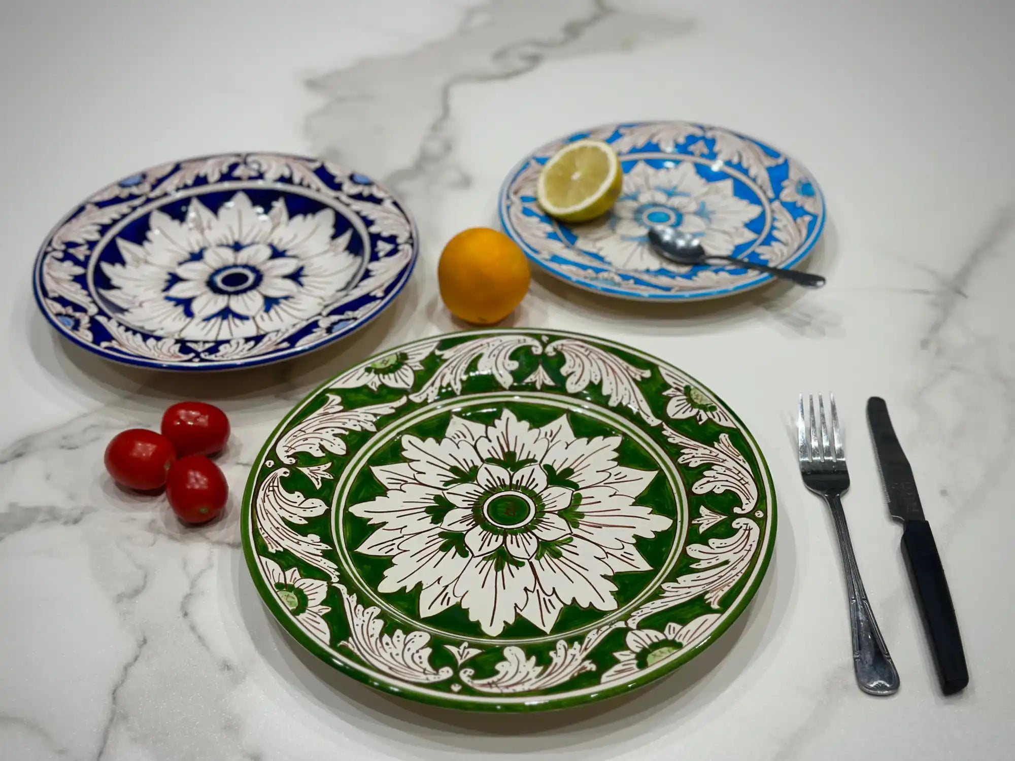 Piatti decorativi con motivi floreali in blu, verde e bianco sono accanto a degli agrumi, pomodori e posate.